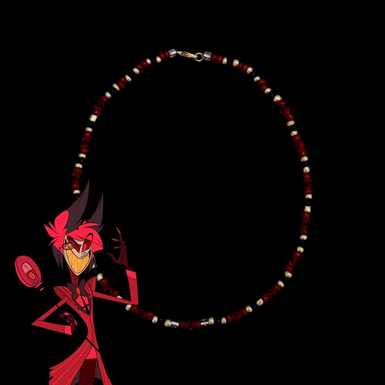 May include: A red and black beaded necklace with a gold clasp. The necklace is circular in shape and is hanging against a black background.