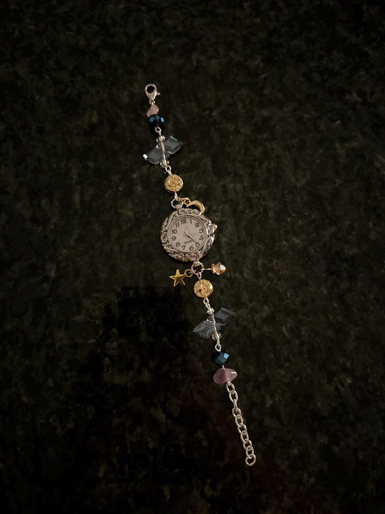 May include: A silver watch with a white face and black Roman numeral markings. The watch is attached to a silver chain with various colored beads, including blue, pink, and gold.