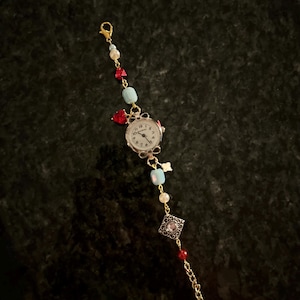 May include: A gold chain bracelet with a small watch face, red and blue beads, and a silver charm with a clear stone.