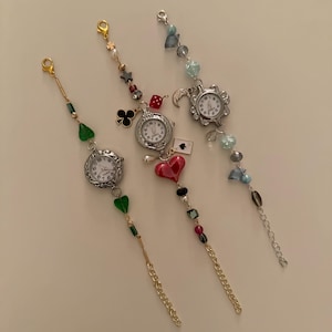 May include: Three silver watch bracelets with different charms. The first bracelet has green and gold beads, the second has red, black, and gold beads, and the third has blue and gold beads.