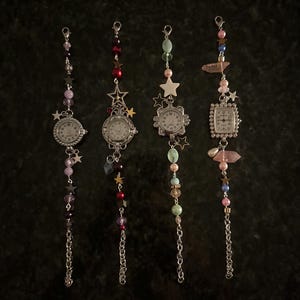 May include: Four vintage-style watch charms with silver chains and decorative beads. Each watch has a white face with black numbers and hands. The charms feature star and gem accents in various colors, including pink, purple, and green.
