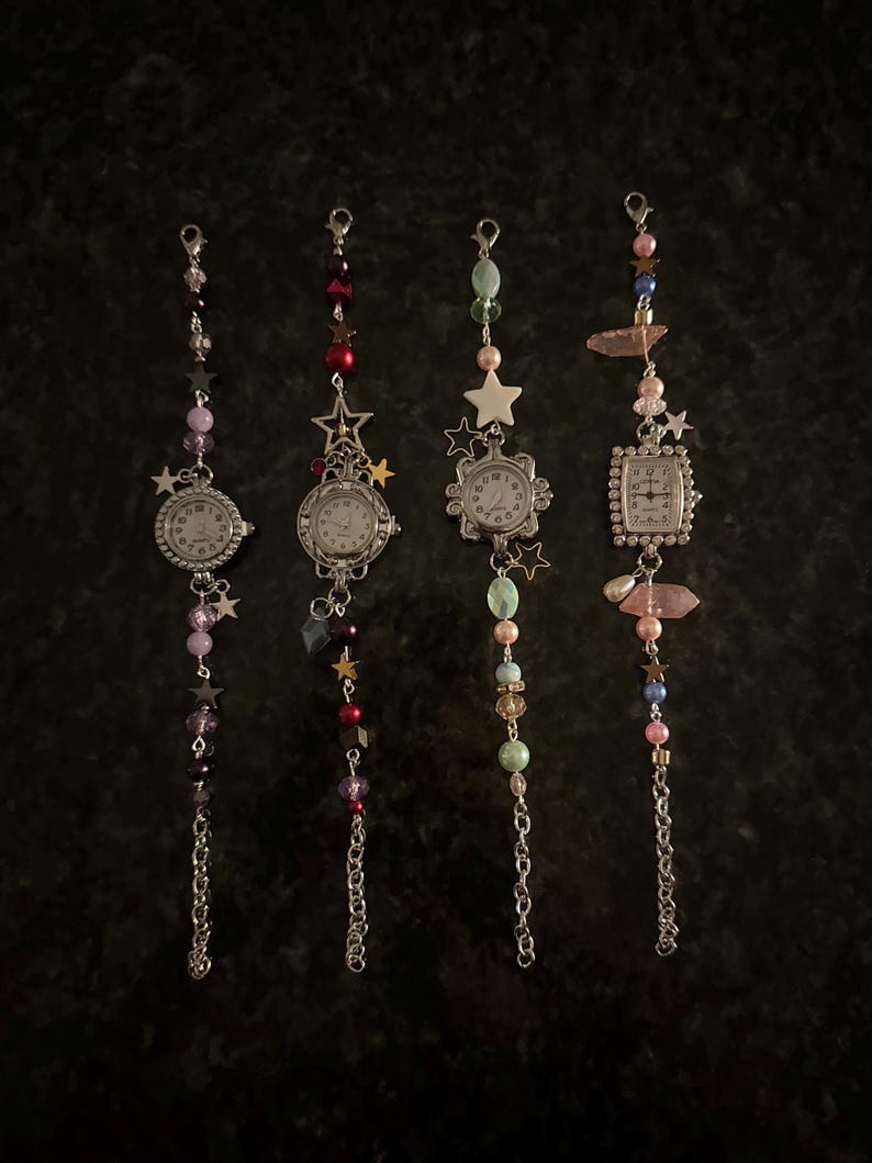 May include: Four vintage-style watch charms with silver chains and decorative beads. Each watch has a white face with black numbers and hands. The charms feature star and gem accents in various colors, including pink, purple, and green.