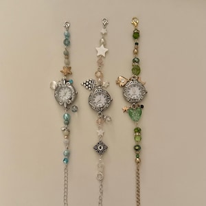 May include: Three decorative wristwatches with silver-toned cases and white faces. Each watch is adorned with beaded chains and charms, including stars, hearts, and sea creatures. The beads are in shades of blue, green, and clear.