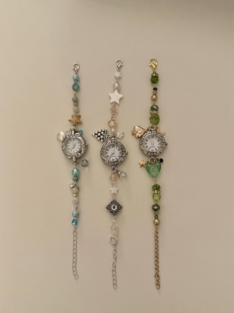 May include: Three decorative wristwatches with silver-toned cases and white faces. Each watch is adorned with beaded chains and charms, including stars, hearts, and sea creatures. The beads are in shades of blue, green, and clear.