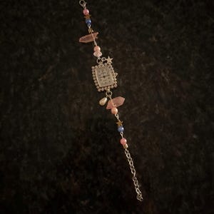 May include: A silver-toned watch bracelet with a rectangular watch face. The bracelet features pink and blue beads, star charms, and pink crystal accents. A small chain extends from the watch face.