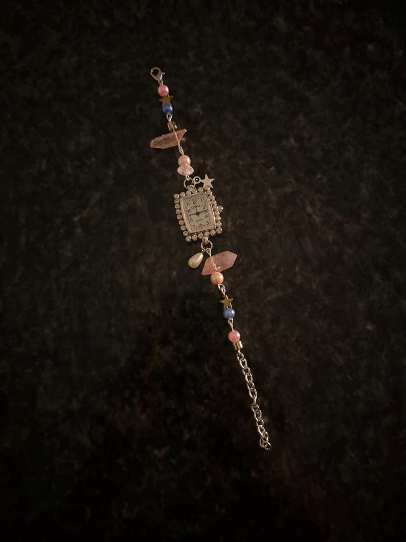 May include: A silver-toned watch bracelet with a rectangular watch face. The bracelet features pink and blue beads, star charms, and pink crystal accents. A small chain extends from the watch face.