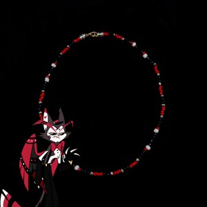 May include: A red, white, and blue beaded necklace with a silver clasp. The necklace is in the shape of a circle and is hanging from a black background. A cartoon character with red and black fur is in the foreground.