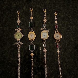 May include: Four gold-tone wristwatches with different coloured charms and crystals hanging from the chain. The watches have white faces with black numbers and hands.