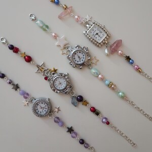 May include: Five beaded charm bracelets with small, round watch faces. The bracelets feature a variety of colorful beads, including pearls, stars, and crystals. The watch faces have silver-colored frames and white dials with black numbers.