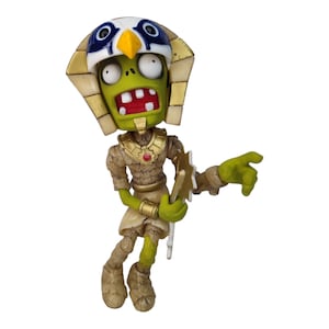 Puede incluir: Una figura de zombi verde que lleva un tocado de faraón dorado y azul y sostiene una espada dorada. El zombi tiene la boca muy abierta y los ojos grandes.