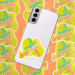 Puede incluir: Un teléfono inteligente blanco con una pegatina en la parte posterior. La pegatina es amarilla, verde y roja con el texto "mello dramatic the original bitter" en ella.