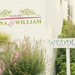 May include: A white wooden sign with the word "WEDDING" written in blue cursive lettering. The sign is propped up in front of a white building with a floral design and the text "ANNA & WILLIAM 03.23.2023" on the wall.