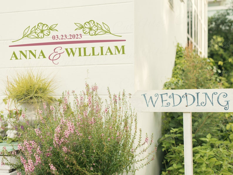 May include: A white wooden sign with the word "WEDDING" written in blue cursive lettering. The sign is propped up in front of a white building with a floral design and the text "ANNA & WILLIAM 03.23.2023" on the wall.