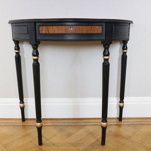 May include: A semi-circular console table featuring a matte black finish with gold-painted accents on the four fluted legs. The central drawer front displays a natural wood grain pattern and is fitted with a small, circular brass-toned metal pull.