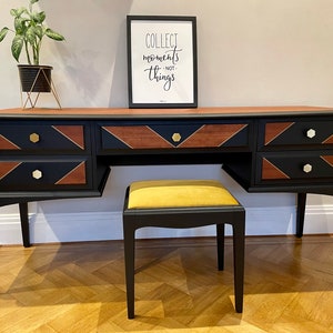 May include: A black wooden vanity desk with a natural wood top and geometric wood inlays on the drawer fronts. The desk features gold hexagonal knobs and is paired with a matching black stool topped with a mustard yellow upholstered seat cushion.
