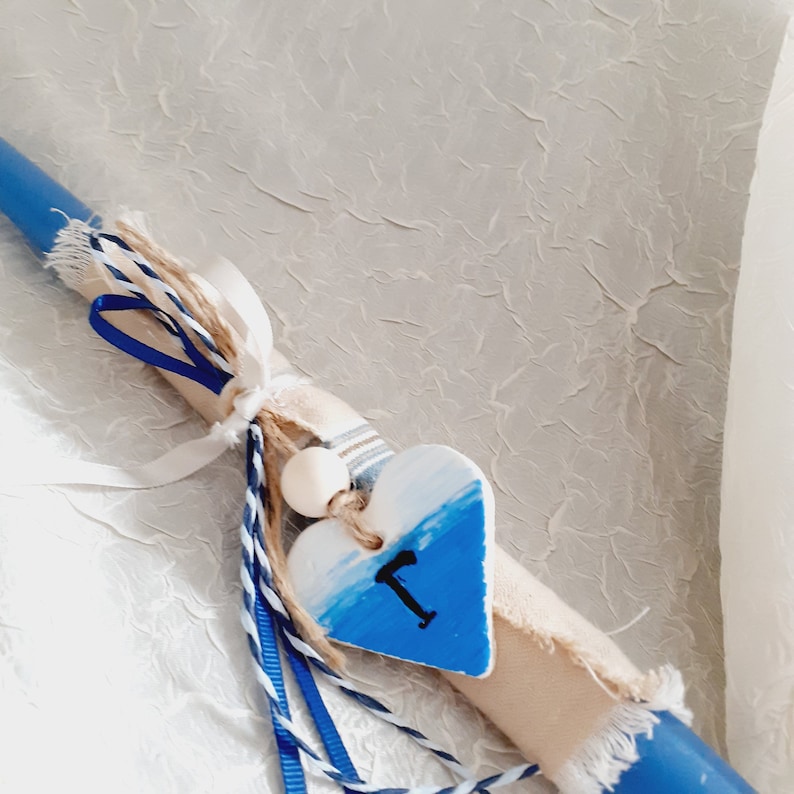 A blue and white Easter lampada candle with a heart-shaped clay ornamen and ribbons attached. The charm is painted with a blue and white ombre design and has the letter "T" painted on it.Lampada is designed in the colours of Greece