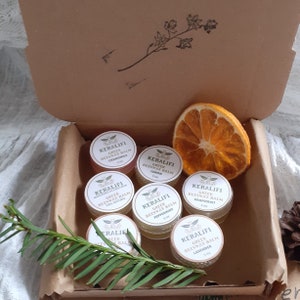 May include: A set of six small glass jars with lids, each containing a different colored balm. The jars are labeled with the text "KERALIFI Greek Beeswax Balm" and the name of a different scent, including chamomile, lemon, grapefruit, peppermint, and lavender. The jars are arranged in a circle on a brown surface.