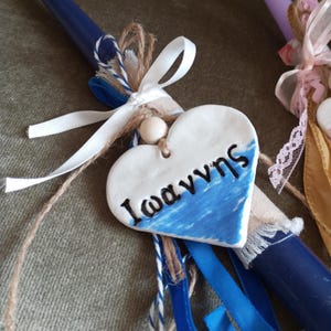 May include: A handmade, heart-shaped ornament with the name "Ιωαννης" in black lettering, painted with blue waves. The ornament is attached to a blue candle with white ribbon, twine, and blue accents. A second candle is visible.