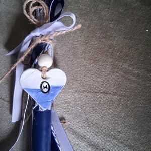 Puede incluir: Una vela azul marino decorada con cintas blancas y azul marino, hilo y un adorno en forma de coraz&oacute;n. El coraz&oacute;n est&aacute; pintado de blanco y azul con un peque&ntilde;o dise&ntilde;o negro. Tambi&eacute;n se adjunta una cuenta de madera.