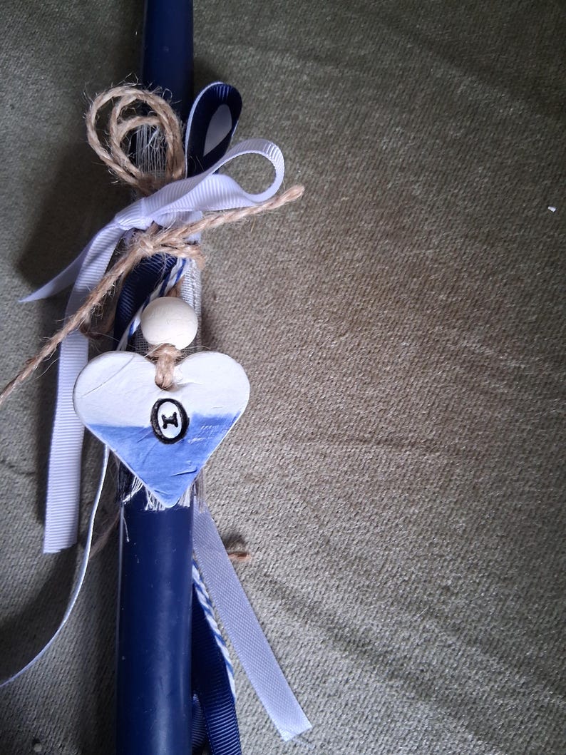 Puede incluir: Una vela azul marino decorada con cintas blancas y azul marino, hilo y un adorno en forma de coraz&oacute;n. El coraz&oacute;n est&aacute; pintado de blanco y azul con un peque&ntilde;o dise&ntilde;o negro. Tambi&eacute;n se adjunta una cuenta de madera.