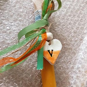 Puede incluir: Una vela amarilla adornada con cintas verdes y naranjas, y un dije en forma de coraz&oacute;n. El dije es blanco con un degradado naranja y la letra "V". La vela est&aacute; envuelta con tela colorida y cuerda.