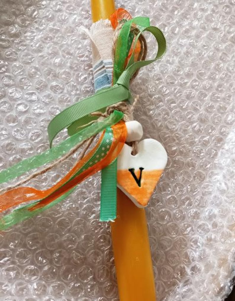 Puede incluir: Una vela amarilla adornada con cintas verdes y naranjas, y un dije en forma de coraz&oacute;n. El dije es blanco con un degradado naranja y la letra "V". La vela est&aacute; envuelta con tela colorida y cuerda.