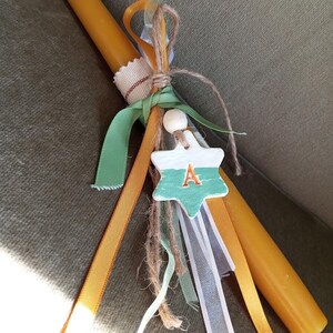 Puede incluir: Una vela decorativa con un dije en forma de estrella. El dije es blanco y verde azulado con la letra "A" en naranja. La vela est&aacute; adornada con cintas verdes, naranjas y blancas, y cuerda.