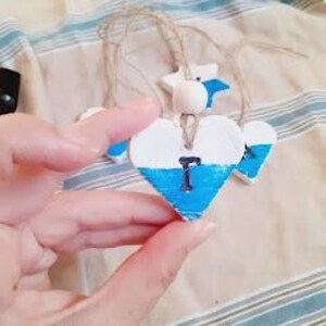 Puede incluir: Adorno artesanal de madera en forma de coraz&oacute;n con un dise&ntilde;o pintado en azul y blanco. El adorno tiene una peque&ntilde;a cuenta de madera y una cuerda para colgar. El coraz&oacute;n est&aacute; sostenido en la mano de una persona. Tambi&eacute;n se ven otros adornos en forma de estrella.
