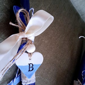 Puede incluir: Una vela azul marino adornada con una cinta de sat&eacute;n blanca, hilo de bramante y un dije en forma de coraz&oacute;n. El dije es blanco y azul con la letra "B". La vela es un objeto decorativo.