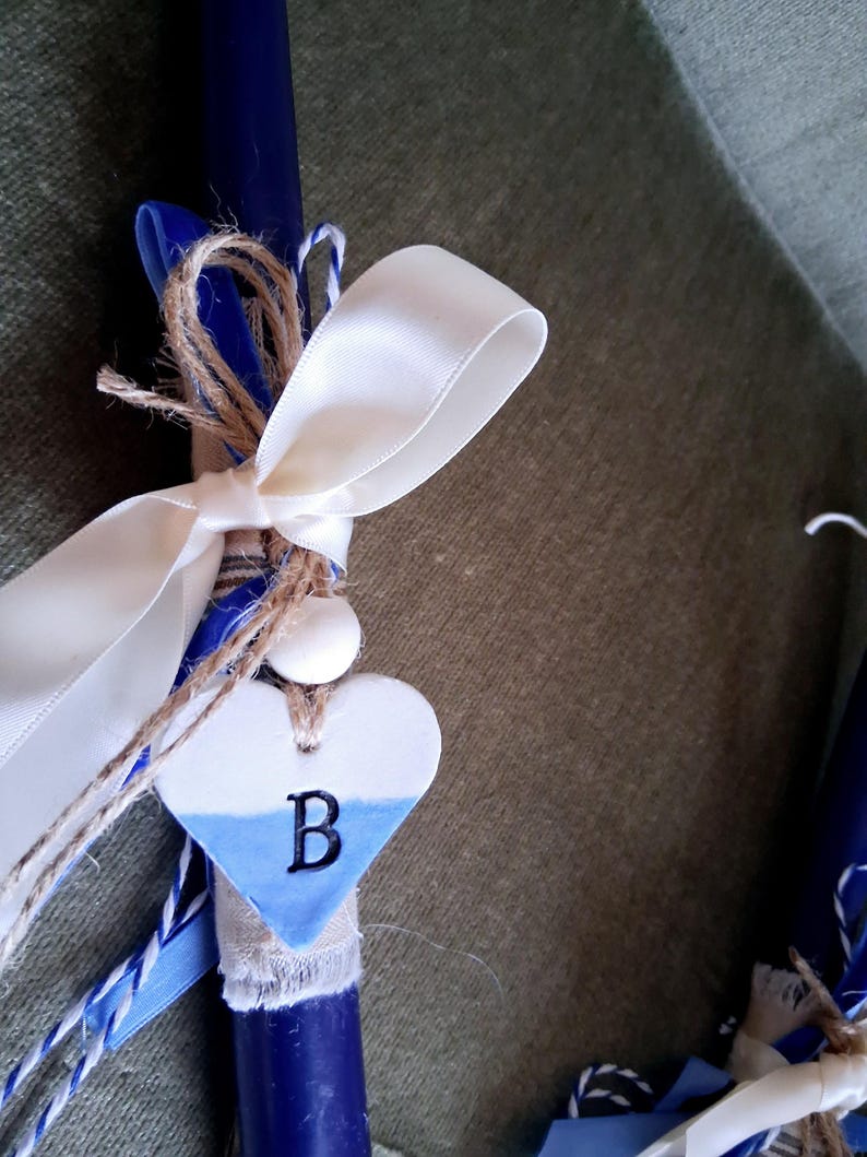 Puede incluir: Una vela azul marino adornada con una cinta de sat&eacute;n blanca, hilo de bramante y un dije en forma de coraz&oacute;n. El dije es blanco y azul con la letra "B". La vela es un objeto decorativo.