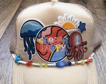 May include: A beige trucker hat featuring a central circular patch with the text "choose happy" in colorful letters. The hat is decorated with embroidered patches of a blue jellyfish, a yellow sun, a blue star, and an octopus, plus a beaded trim.