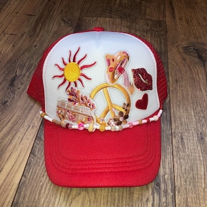 May include: A red and white trucker hat featuring embroidered patches of a yellow sun, a mushroom, a peace sign, a red glittery pair of lips, a small red heart, and a camper van. A string of multicoloured beads is attached across the front brim.