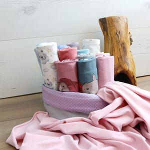 Könnte beinhalten: Ein Korb gefüllt mit einer Vielzahl von bunten Baby-Swaddles und Decken. Die Swaddles haben verschiedene Muster und Designs, darunter Sterne, Wolken und Tiere. Der Korb ist mit einem hellvioletten Stoff mit weißen Punkten ausgekleidet.
