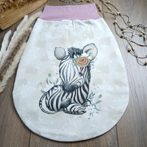 Könnte beinhalten: Ein weißer Schlafsack mit rosa Besatz und einem niedlichen Zebra-Druck. Das Zebra trägt eine Blumenkrone und hat einen weißen Hintergrund mit einem Blumenmuster.