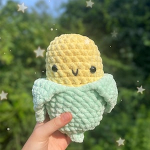Op de afbeelding: Een gehaakt knuffel maïs met een groene schil en een gele kolf. De maïs heeft een glimlachend gezicht met zwarte ogen en een kleine mond.