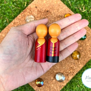 Puede incluir: Dos muñecos de madera con ropa pintada de rojo y dorado. Los muñecos están de pie sobre una mano.