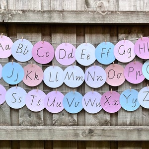 以下が含まれることがあります： アルファベットの各文字が円形に書かれたカラフルなアルファベットバナー。円はピンク、ブルー、パープルの色合いで、白い紐で繋げられています。
