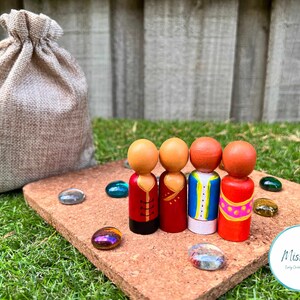 Könnte beinhalten: Vier Holz-Steckpuppen, die in farbenfrohe Kleidung gekleidet sind, stehen auf einem Korkbrett mit verstreuten Glasperlen. Im Hintergrund befindet sich ein Jutesack. Die Puppen tragen rote, blaue und gelbe Kleidung mit aufwendigen Details.