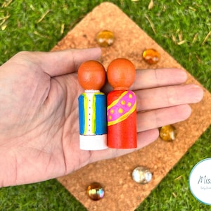 May include: Two wooden peg dolls with orange heads. One doll wears a blue and white outfit, the other a red outfit with a pink and yellow design. The dolls are on a brown surface with decorative stones.