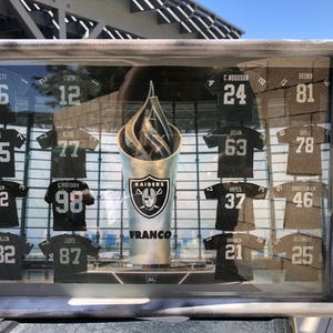 May include: A wooden tray with a glass insert displaying a collection of black and brown football jerseys with numbers and names. The jerseys are arranged in a grid pattern and are surrounded by a silver frame. The tray also features a silver trophy with the word "RAIDERS" and the name "FRANCO" printed on it.