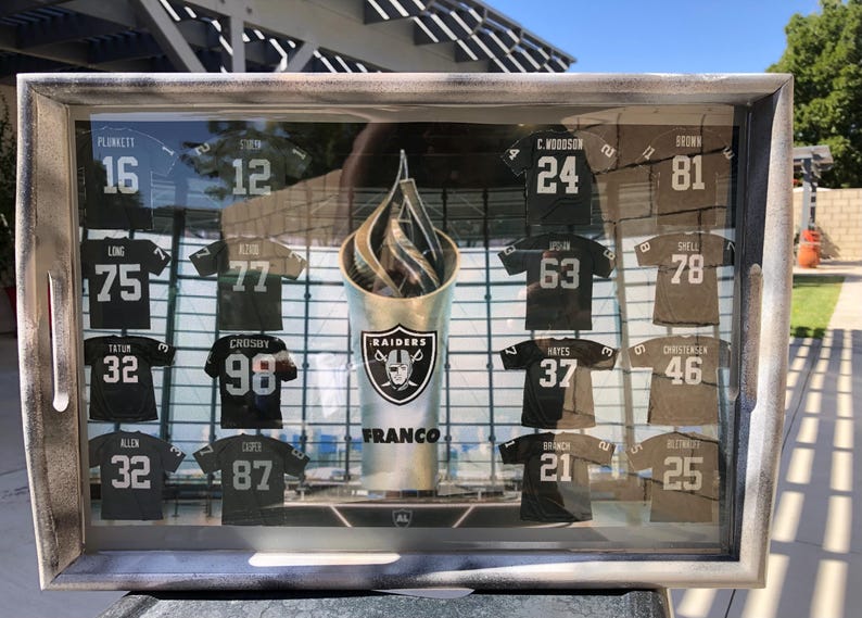May include: A wooden tray with a glass insert displaying a collection of black and brown football jerseys with numbers and names. The jerseys are arranged in a grid pattern and are surrounded by a silver frame. The tray also features a silver trophy with the word "RAIDERS" and the name "FRANCO" printed on it.