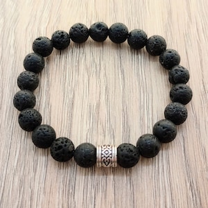 May include: A black lava rock beaded bracelet with a silver charm. The bracelet is made of natural lava rock beads and has a silver charm with a geometric design.