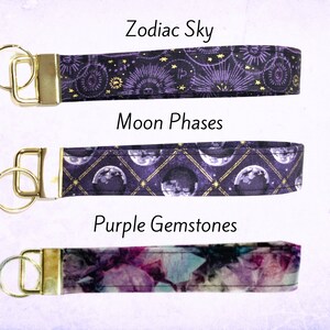 May include: Three purple and gold keychains with different designs. The top keychain features a zodiac sky design, the middle keychain features a moon phases design, and the bottom keychain features a purple gemstones design.