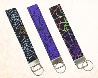 May include: Three Halloween-themed wrist keychains. The keychains feature a black, purple, and black fabric with spiderweb designs. Each keychain has a silver metal ring for attaching keys.
