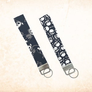 May include: Two black keychains with silver key rings. One keychain has a white skeleton cat design and the other has a white skull and crossbones design.