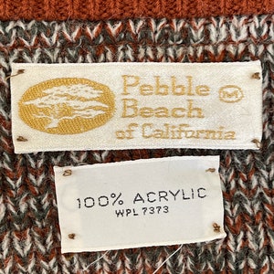 May include: Close-up of a sweater with a textured pattern in shades of brown, white, and gray. The top portion is a solid rust color. Two tags read "Pebble Beach of California" and "100% Acrylic".