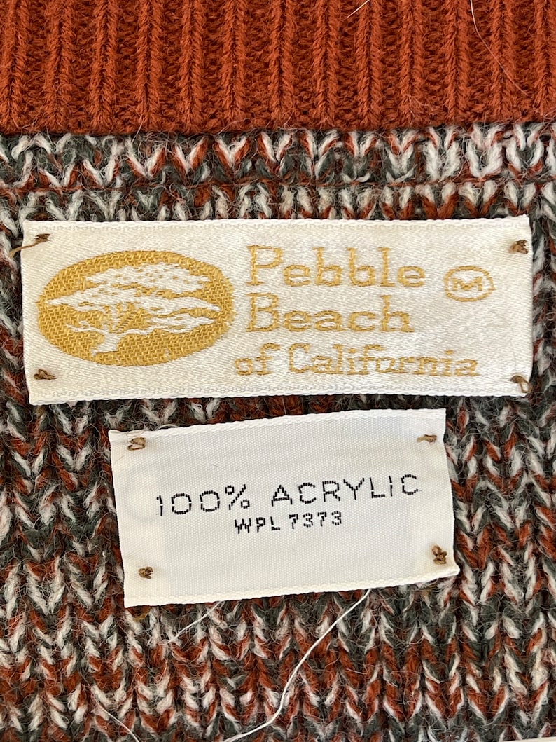 May include: Close-up of a sweater with a textured pattern in shades of brown, white, and gray. The top portion is a solid rust color. Two tags read "Pebble Beach of California" and "100% Acrylic".