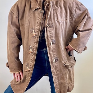 May include: Brown coat with toggle closures and front pockets. The coat has a relaxed fit, frayed edges, and a button-down collar. The coat is worn over a black top and blue jeans.