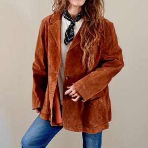 May include: A rust-colored suede blazer featuring a notched lapel, two front buttons, and two patch pockets. The blazer is worn over a white t-shirt and a black and white patterned neck scarf, paired with blue denim jeans.