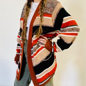 May include: A multi-colored open cardigan with a striped pattern in shades of brown, black, orange, and cream. The cardigan has long sleeves and a brown trim. The garment is made of a thick knit material.